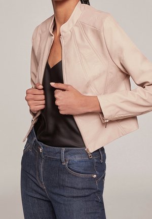 Personne portant une veste en cuir beige clair sur un top noir et un jean bleu, tenant la fermeture éclair de la veste avec les deux mains.