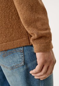 Hnědý texturovaný svetr s těsným střihem, doplněný světle modrými džíny. Viditelné švy na zadní kapse džínů.