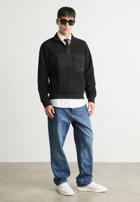 Felpa nera con colletto zip, tasca frontale, indossata sopra una camicia bianca con colletto, abbinata a jeans blu e sneakers bianche.