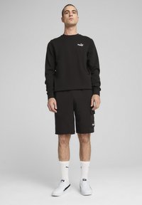 Black sweatshirt with crew neck and logo, paired with black shorts featuring side pocket. Worn with white socks and sneakers.
