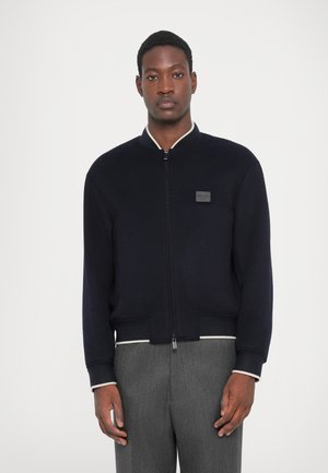 Navy zip-up bomber jacket with contrasting white ribbed collar and cuffs, featuring a small logo patch on the chest. Soft knit texture.