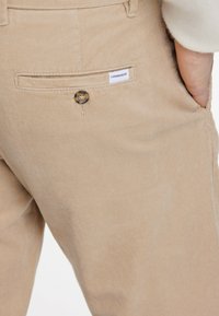 Beige corduroy pants with a visible back pocket, featuring a button closure and a small label that reads "LINDHBERGH." Textured fabric detail.