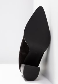 Zapato de ante negro con punta afilada y ribete de tachuelas a lo largo de la suela, con un tacón cuadrado, apoyado en un fondo blanco en esquina.