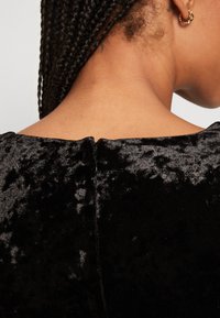 Robe en velours noir avec un décolleté large, comportant une fermeture éclair discrète à l'arrière. Le tissu a une texture lisse et réfléchissante.