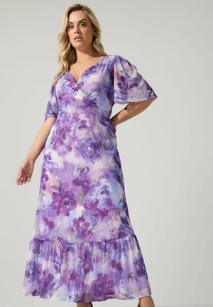 Frau in einem lila und lavendelfarbenen Wickelkleid mit Blumenmuster, Puffärmeln und Volant am Saum, stehend vor einem schlichten Hintergrund.