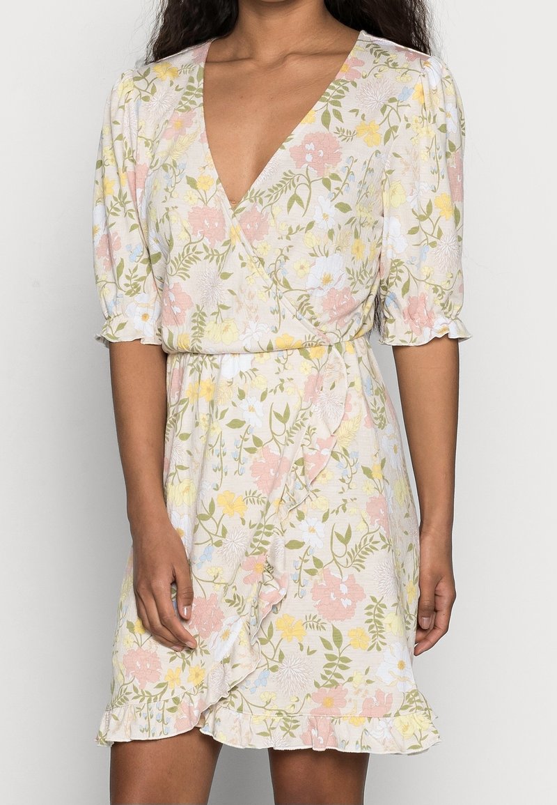 Robe portefeuille fleurie avec un décolleté en V, des manches courtes bouffantes et un ourlet à volants. Le tissu est léger avec des motifs pastel rose, jaune et vert.