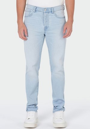Mann trägt hellblaue Slim-Fit-Jeans und weiße Sneaker und steht vor einem schlichten Hintergrund.