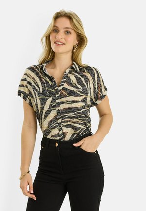 Jeune femme souriante, portant une chemise à manches courtes à imprimé animal boutonnée et rentrée dans un pantalon noir taille haute, la main dans la poche.