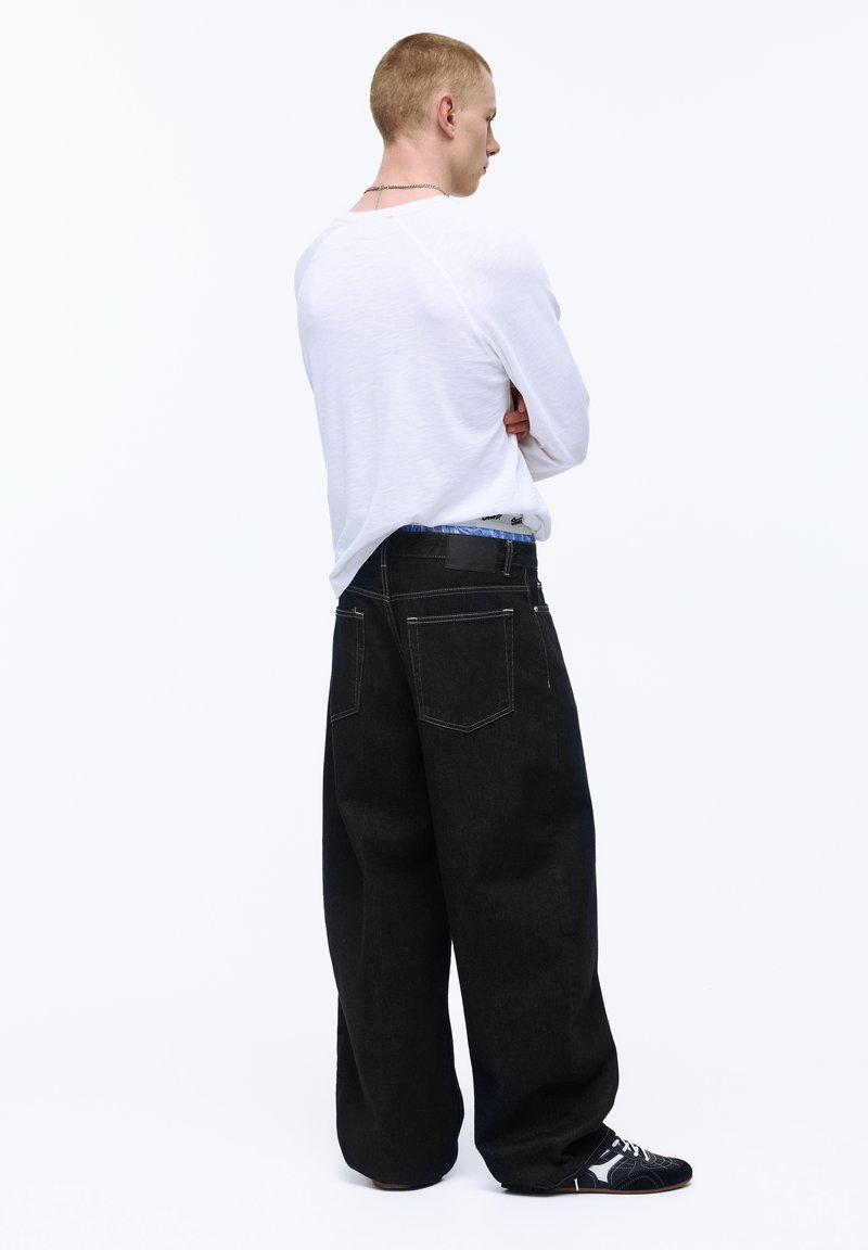 Jeune homme aux cheveux blonds courts, portant une chemise blanche à manches longues, un large jean noir et des baskets noires, debout les bras croisés, de profil.