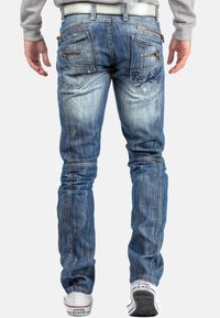 Jean en denim bleu délavé avec une coupe slim. Présente plusieurs poches zippées et des détails brodés à l'arrière. Porté avec des baskets blanches.