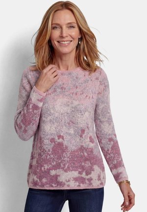 Femme aux cheveux blonds mi-longs souriante, portant un pull à manches longues à motif rose et violet et un jean bleu foncé.