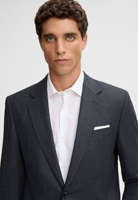 Marineblauwe geruite blazer met notch revers, enkele knoopsluiting, pochet en gelaagd over een witte dressshirt met knoopsluiting.