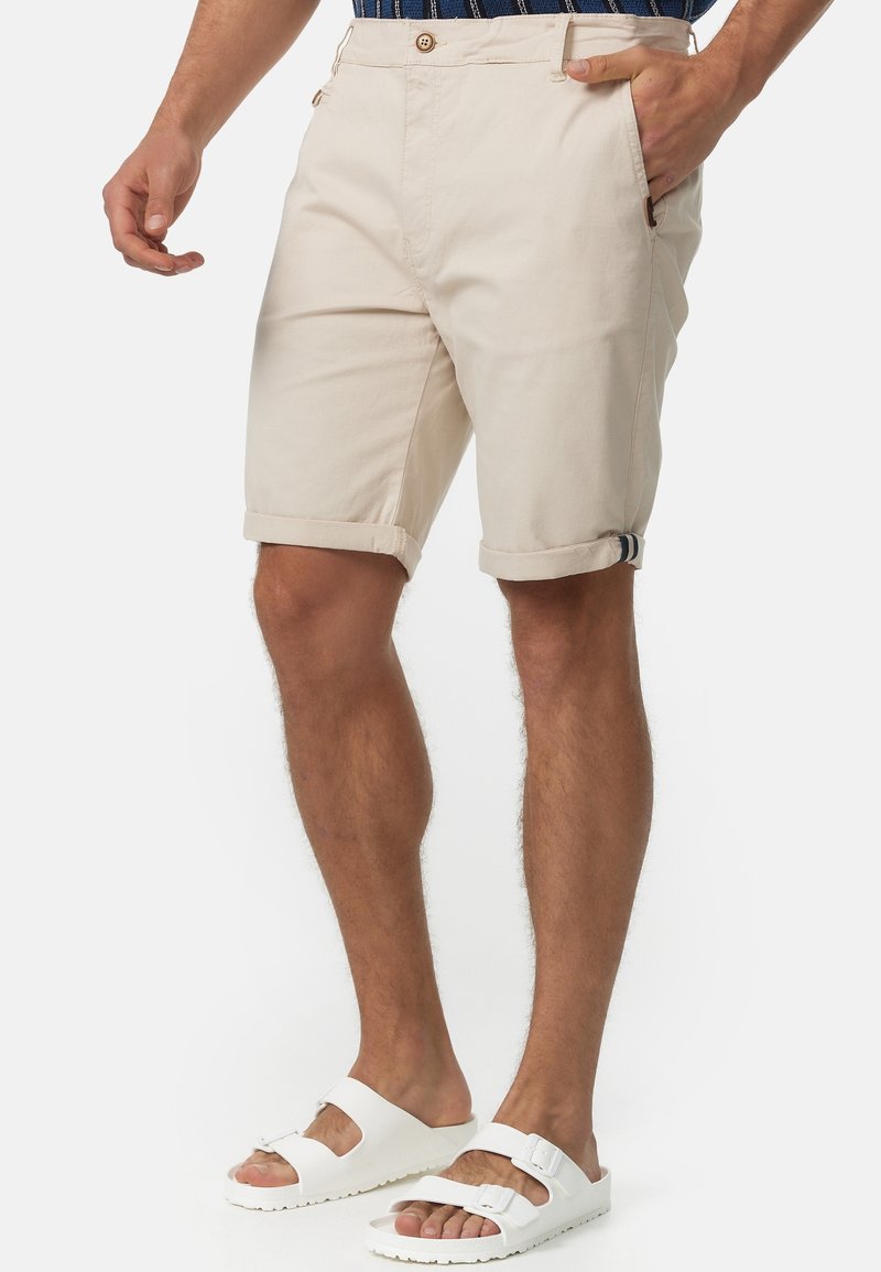 Homme portant un short beige retroussé, une chemise rayée bleu marine et des sandales blanches à boucle sur un fond uni.
