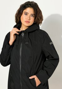 Chaqueta impermeable negra con capucha, cierre de cremallera y toggles ajustables. Se muestra con una mano en el bolsillo y textura suave.