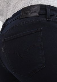 Acercamiento a un bolsillo trasero y la cinturilla de unos vaqueros Levi's de un azul oscuro, con un parche de cuero de Levi Strauss & Co. en una persona que lleva una camiseta gris.
