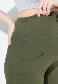 Legíny olivové barvy vyrobené z měkké, elastické látky mají boční kapsy a přiléhavý střih s kontrastními švy.