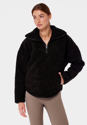 Młoda kobieta z włosami spiętymi do tyłu, ubrana w czarny polar z zamkiem przy kołnierzu i beżowe legginsy, pozująca z jedną ręką w kieszeni.