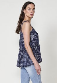 Marineblauwe mouwloze top met een geraffineerde taille, voorzien van een geweven patroon en verstelbare strikbandjes. Gecombineerd met lichtblauwe spijkerbroek.