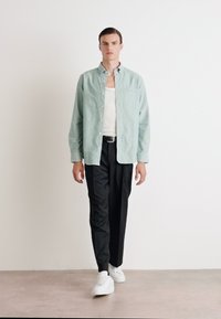 Camicia verde chiaro con bottoni sopra una canotta bianca, abbinata a pantaloni neri e sneaker bianche. Design minimalista, texture liscia.