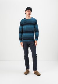 Strikket blå og mørk stripet genser, båret med mørke bukser og brune slip-on sko; modellen har også på seg en svart caps.