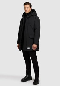 Black insulated parka with a hood, featuring a front zipper, two side pockets, and leather accents, worn over a black turtleneck and pants.