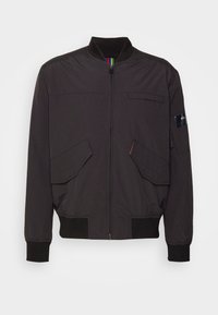Veste bomber noire avec fermeture éclair frontale, deux grandes poches à rabat inclinées, poignets et taille côtelés, et un petit patch sur la manche gauche.