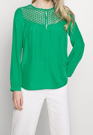 Zelená halenka s krajkovým sedlem, dlouhými rukávy a zapínáním na knoflíky. Vyrobená z hladké látky, je kombinována s bílými kalhotami.