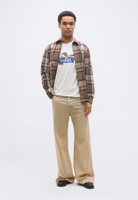 Άνδρας που φοράει μπεζ φαρδιά παντελόνια, λευκό μπλουζάκι με σχέδιο ψαριού, καφέ πουκάμισο με καρό σχέδιο ανοιχτό και μαύρα παπούτσια, στέκεται μπροστά σε λευκό φόντο.