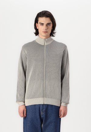 Grijze ribbelige zip-up jas met hoge kraag, textuurstof, lange mouwen en contrasterende rand. Draagt met blauwe denim broek.