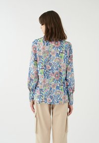 Blusa a maniche lunghe floreale con base blu. Presenta vivaci motivi rosa, verdi e viola. Tessuto morbido, vestibilità comoda e polsini con bottoni.