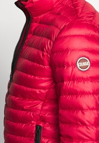 Červená péřová bunda s prošívaným designem, vysokým límcem, černým zipem a kulatou nášivkou na horním rukávu.