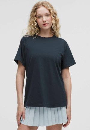 Donna con capelli biondi ricci che indossa una semplice t-shirt nera a maniche corte e una gonna plissettata azzurra, in piedi davanti a uno sfondo uniforme.