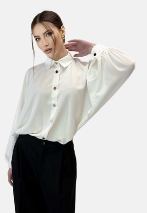 Blusa blanca de botones con cuello, mangas largas fluidas y botones dorados, combinada con pantalones negros de tiro alto. Textura de tela suave.