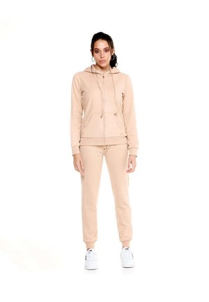 Une femme se tient devant un fond blanc, vêtue d’un sweat à capuche beige zippé, d’un pantalon de jogging assorti et de baskets blanches.