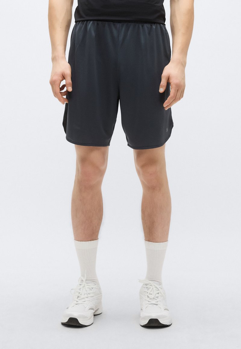 Pantaloni scurți atletici negri cu talie elastică, buzunare laterale și un material neted, ușor. Purtate cu adidași și șosete albe.