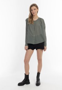 Camicetta verde con colletto rotondo e maniche lunghe, abbinata a pantaloni corti di jeans neri e stivali neri alla caviglia. Tessuto dalla consistenza liscia.