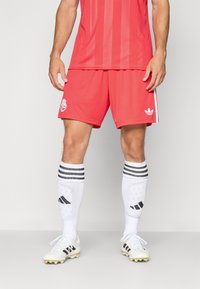 Rode voetbalshirt met verticale strepen, bijpassende rode shorts, witte kniekousen met zwarte strepen en witte voetbalschoenen met zwarte accenten.
