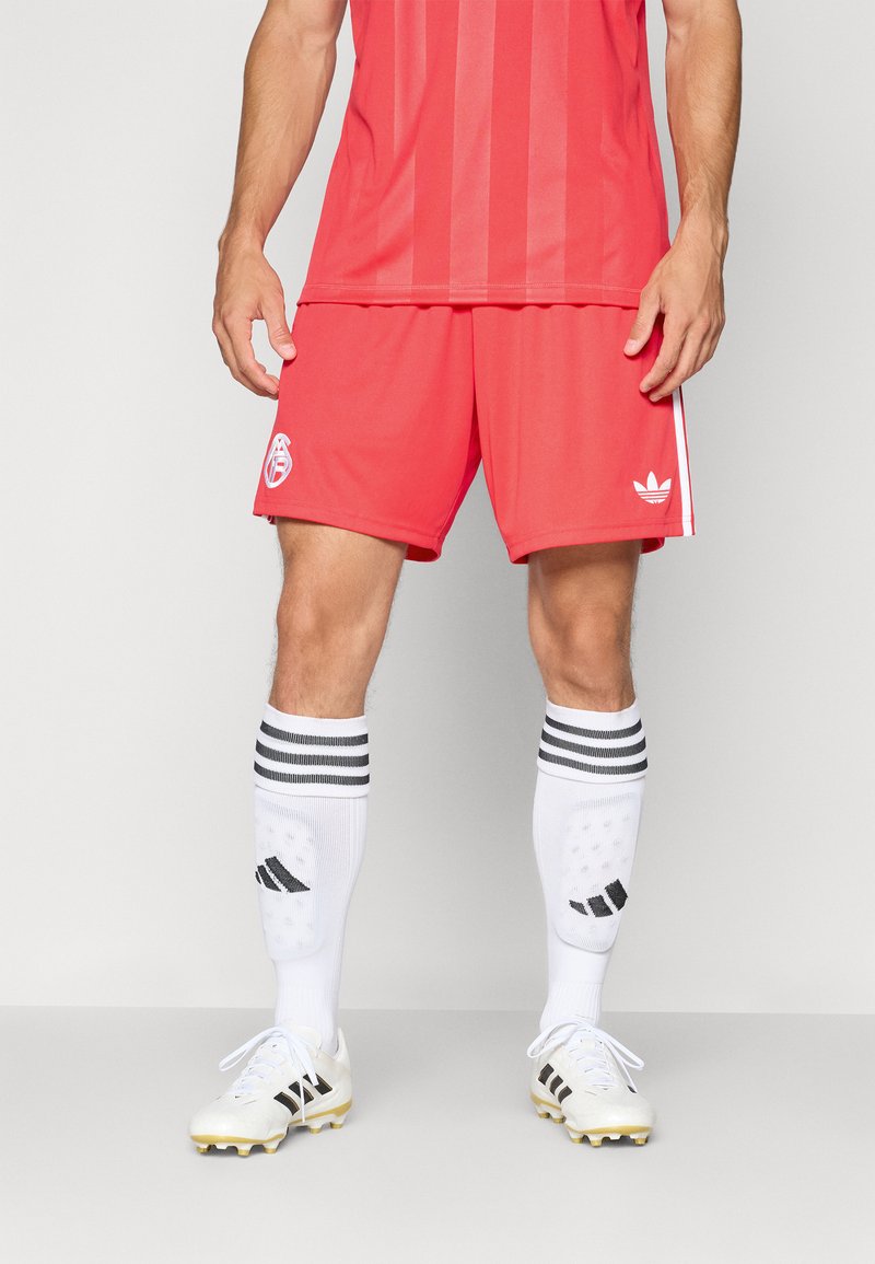 Rode voetbalshirt met verticale strepen, bijpassende rode shorts, witte kniekousen met zwarte strepen en witte voetbalschoenen met zwarte accenten.