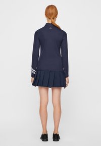 Mörkblå långärmad topp med logotyp på baksidan, kombinerad med en mörkblå veckad kjol. Materialet verkar slätt; skorna är svarta sneakers.