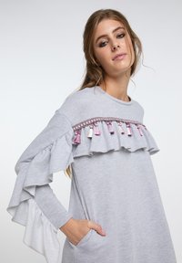 Graues, langärmliges Kleid mit einem rüschartigen Akzent am Ausschnitt, ausgestattet mit dekorativen Quasten in Pink und Weiß. Mit seitlichen Taschen.