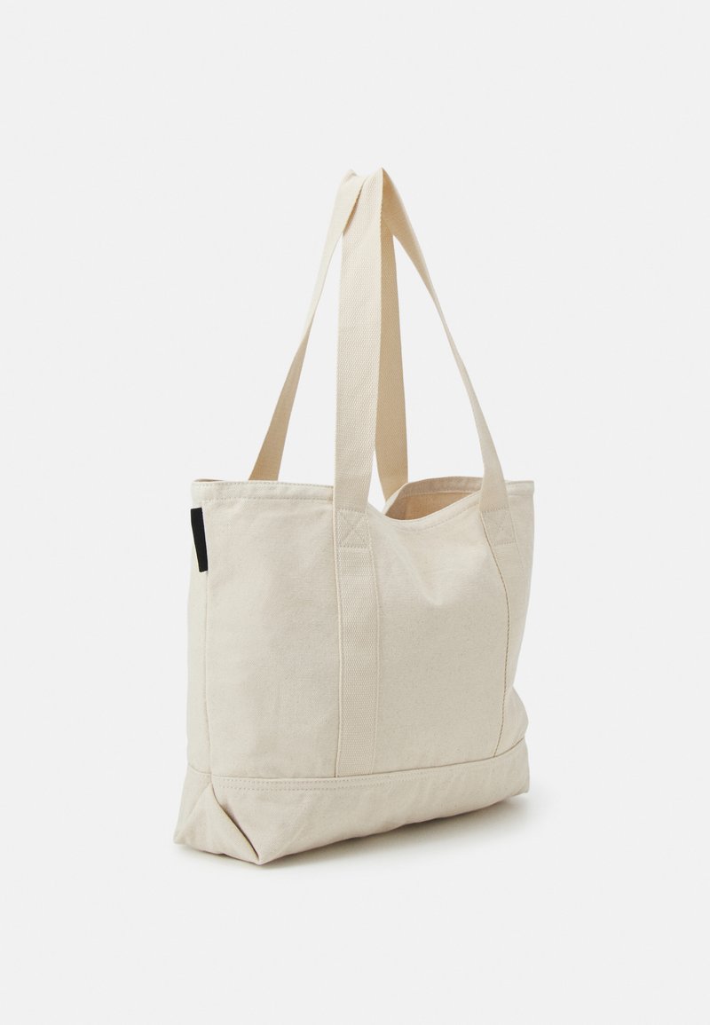 Bolsa de lona beige con asas largas, de forma rectangular, con costura doble y una etiqueta lateral. Textura suave con una base estructurada.