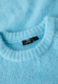 Pull tricoté doux bleu clair avec col côtelé et étiquette de vêtement noire portant l'inscription "LMTD" cousue à l'intérieur de l'encolure.