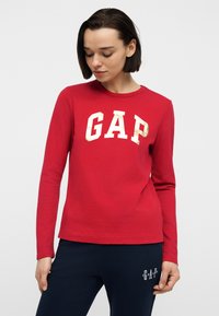 Γυναίκα με σκούρα κοντά μαλλιά, φοράει κόκκινο μακρυμάνικο πουκάμισο με χρυσό λογότυπο "GAP" και μπλε παντελόνι με λευκό κείμενο "GAP" στο μηρό.