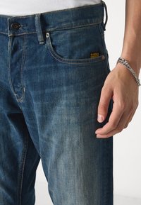 Donkerblauwe spijkerbroek met contrasterende stiksels, een knoopsluiting en een logo-label op de voorzak. Hand rustend nabij de taille.