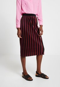 Gonna nera con strisce verticali multicolori, lunghezza fino al ginocchio, caratterizzata da un design a pieghe e una vita elastica. Abbinata a una camicia button-up rosa e sandali con borchie.