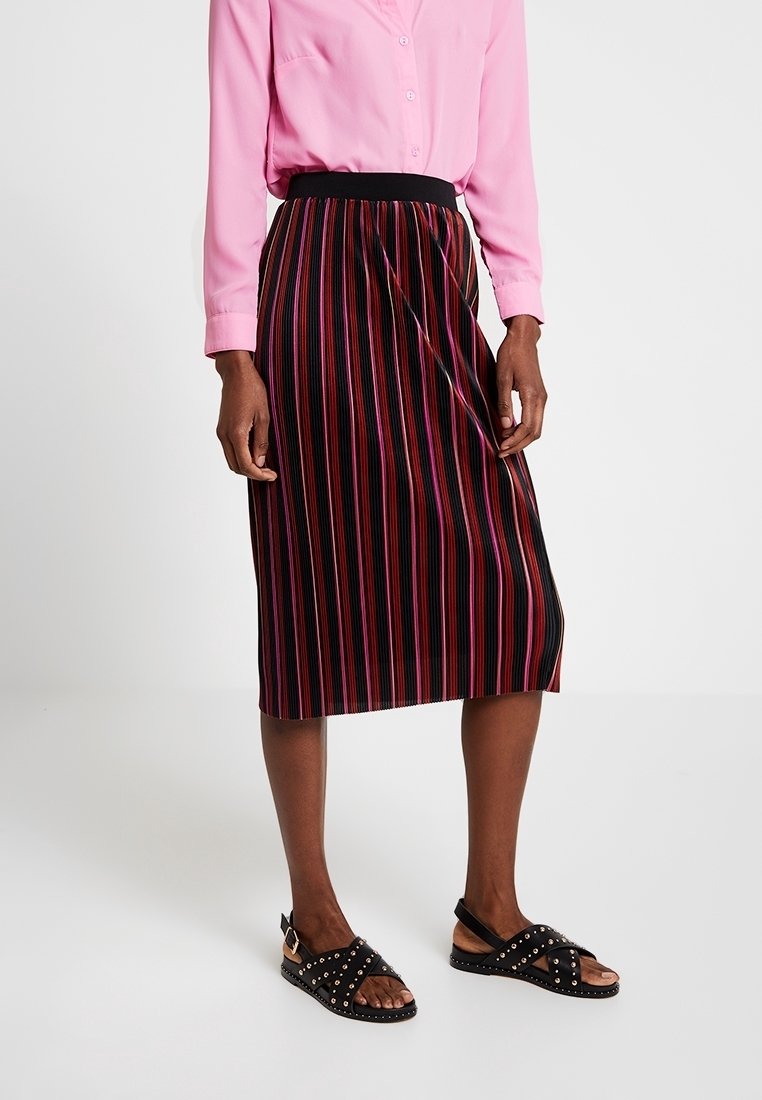 Gonna nera con strisce verticali multicolori, lunghezza fino al ginocchio, caratterizzata da un design a pieghe e una vita elastica. Abbinata a una camicia button-up rosa e sandali con borchie.