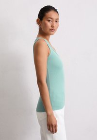Débardeur léger bleu clair à côtes avec des bretelles fines, design ajusté et texture lisse, associé à un pantalon blanc. Le modèle se tient de profil.