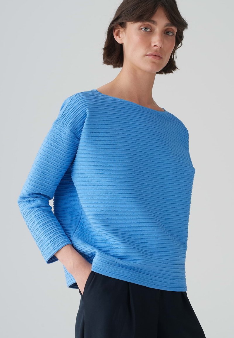 Blauwe textuur trui met horizontale ribbels, ronde halslijn en lange mouwen, gecombineerd met zwarte broek. Glad stofafwerking.