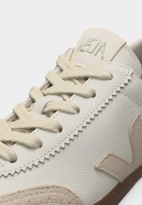 Κοντινό πλάνο κρεμ αθλητικού παπουτσιού Veja με δερμάτινη υφή, μπεζ κορδόνια, ζιγκ ζαγκ περιθώριο, σουέντ λεπτομέρειες και καφέ καουτσούκ σολα.