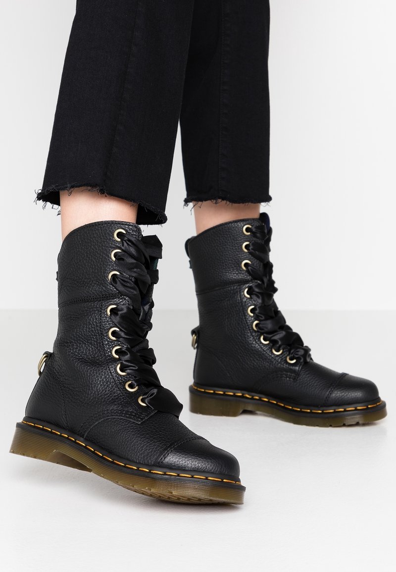 Bottes montantes en cuir noir avec semelles épaisses translucides et coutures jaunes, portées avec un pantalon court noir effiloché sur un sol blanc.